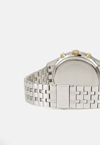 Reloj de acero inoxidable plateado con pulsera trenzada, caja redonda, acentos dorados en los pulsadores y respaldo liso y mate.