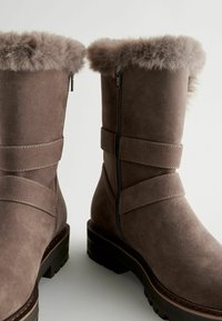 Brown suede boots with a faux fur trim and zip closure. Features two wide straps around the shaft and a textured outsole for grip.