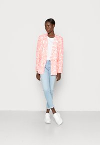 Blazer com padrão floral rosa sobre uma t-shirt branca, combinado com jeans azul claro e sapatilhas brancas. O tecido parece texturizado e leve.