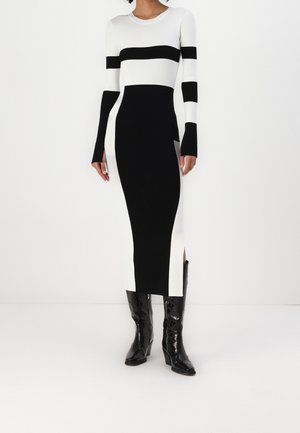 Mujer vestida con un vestido largo bicolor en blanco y negro, de mangas largas y botas negras hasta la rodilla, de pie frente a un fondo blanco.