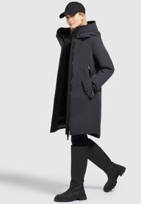 Black long coat with an oversized hood, zippered pockets, and a smooth texture, paired with knee-high black boots. Set against a plain background.
