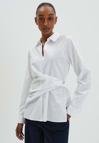 Weißes Button-Up-Hemd mit taillierter Passform, das ein gedrehtes Vorderdesign und silberne Hardware-Akzente an der Seite aufweist. Lange Ärmel, glatte Textur.