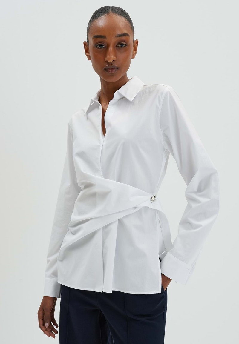 Weißes Button-Up-Hemd mit taillierter Passform, das ein gedrehtes Vorderdesign und silberne Hardware-Akzente an der Seite aufweist. Lange Ärmel, glatte Textur.