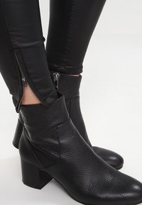 Bottines en cuir noir texturé avec des talons carrés, portées avec un pantalon noir doté de zips latéraux au niveau des chevilles.