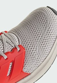 Chaussure de sport légère avec une tige en mesh gris, des accents en plastique rouge et des lacets gris. Surface texturée et semelle rembourrée visibles.