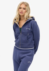 Felpa con zip di colore blu navy e design cropped, con tasche frontali e ricamo del logo bianco; abbinata a pantaloni della tuta coordinati.