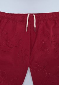 Rode korte broek met een gestructureerde stof en een elastische tailleband, voorzien van witte koordjes en bladmotieven in de stof gedrukt.