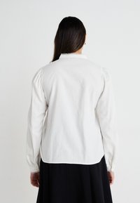 Chemise blanche à manches longues en coton, avec des manches ballon et un col en dentelle. Texture subtile avec des détails de boutons sur les manches.