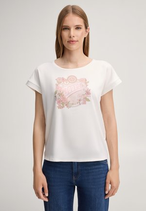Vrouw met steil lichtbruin haar draagt een wit T-shirt met bloemenprint en ‘Joop!’-tekst en een blauwe spijkerbroek, staand tegen een effen achtergrond.