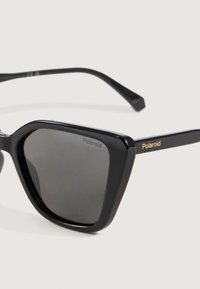 Mustat suorakulmaiset Polaroid -aurinkolasit tummilla linsseillä ja kultaisella logolla temppeleissä, yksinkertaista valkoista taustaa vasten.