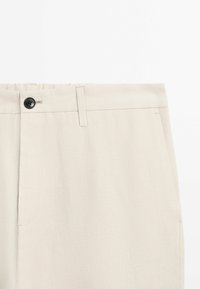 Section de la taille d’un pantalon beige avec bouton noir, passants de ceinture et poche latérale visibles sur fond blanc.