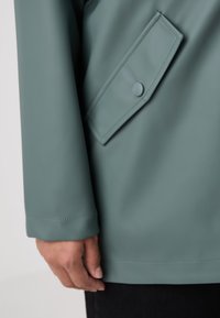 Een groene waterdichte jas met een glad oppervlak, voorzien van een diagonale klepzak die met een knoop is bevestigd, weergegeven vanaf de elleboog naar beneden.