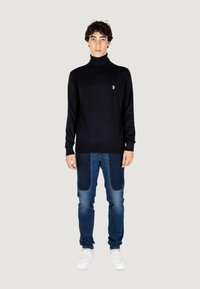 Maglione a collo alto nero con logo dorato, abbinato a jeans blu con pannelli in tessuto a contrasto e sneakers bianche.