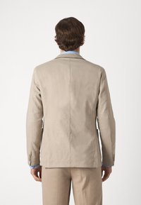 Giacca beige con una vestibilità sartoriale, fessura centrale e chiusura a due bottoni. Presenta una texture liscia e una camicia azzurro chiaro in contrasto sotto.