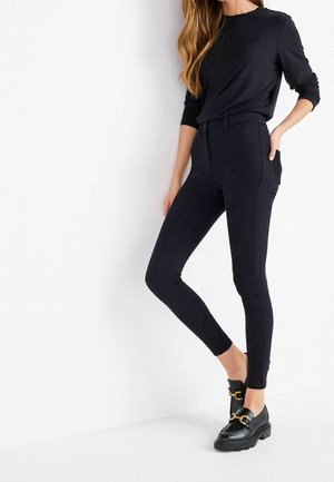 Femme portant un pantalon noir ajusté, un haut noir à manches longues et des mocassins noirs avec des boucles dorées, debout devant un fond blanc uni.