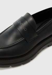 Black leather loafers with a raised logo band across the upper. Features a sleek design and textured stitching, complemented by a thick sole.