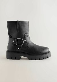 Bottine en cuir noir avec une semelle épaisse, dotée d'une bride avec un détail métallique circulaire et d'une tige texturée.