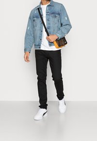 Man som bär en ljusblå denimjacka, vit t-shirt, svarta skinny jeans, vita sneakers och bär en svart och orange axelremsväska.