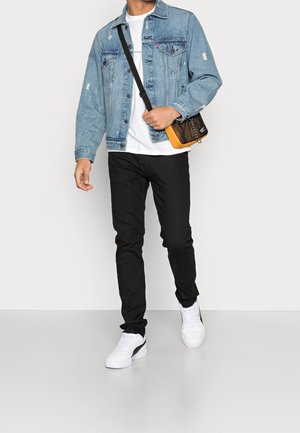 Uomo che indossa una giacca di jeans azzurro chiaro, una t-shirt bianca, jeans skinny neri, sneakers bianche e porta una borsa a tracolla nera e arancione.