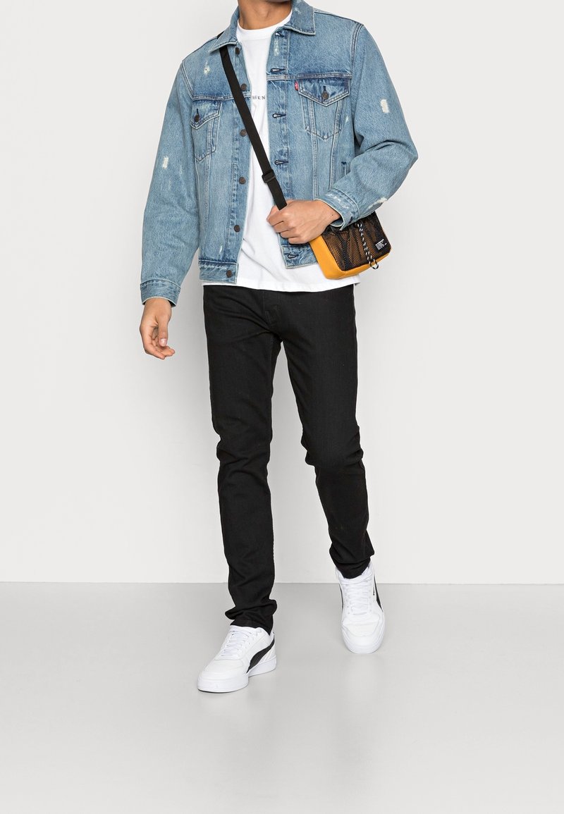 Man som bär en ljusblå denimjacka, vit t-shirt, svarta skinny jeans, vita sneakers och bär en svart och orange axelremsväska.