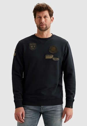 Muž v čiernej mikine s dlhým rukávom a viacerými vyšívanými nášivkami, v svetlomodrých džínsoch, stojaci pred jednoduchým sivým pozadím.