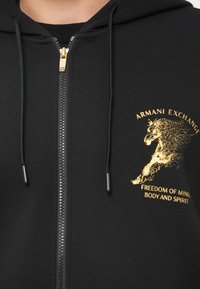 Juodas zip-up džemperis su auksiniu arklių grafikų ir užrašu "ARMANI EXCHANGE" bei "LAISVĖ PROTUI, KŪNUI IR DVASIAI" ant krūtinės.