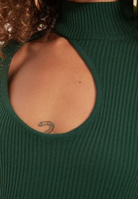 Haut vert à col montant à nervures avec une découpe en goutte d'eau au niveau de la poitrine, révélant un petit tatouage de serpent. Le tissu a un aspect texturé.