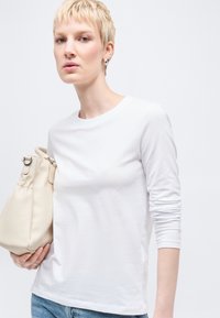 Γυναίκα με κοντά ξανθά μαλλιά που φοράει λευκό μακρυμάνικο πουκάμισο και τζιν, κρατώντας μια μπεζ τσάντα χειρός σε απλό ανοιχτό φόντο.