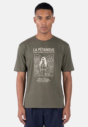 Uomo che indossa una maglietta verde oliva con grafica di donna che gioca a pétanque e testo “La Pétanque non è uno sport, è uno stile di vita.”