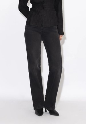Persona che indossa jeans neri a vita alta, stivali neri con tacco e punta a punta, e un top nero a maniche lunghe, in piedi davanti a uno sfondo bianco.