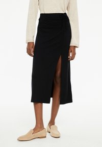 Falda midi negra de tela suave con una abertura frontal. Cuenta con una cintura con cordón y una textura suave, combinada con zapatos de lona ligeros en color beige claro.