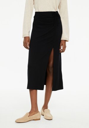 Falda midi negra de tela suave con una abertura frontal. Cuenta con una cintura con cordón y una textura suave, combinada con zapatos de lona ligeros en color beige claro.