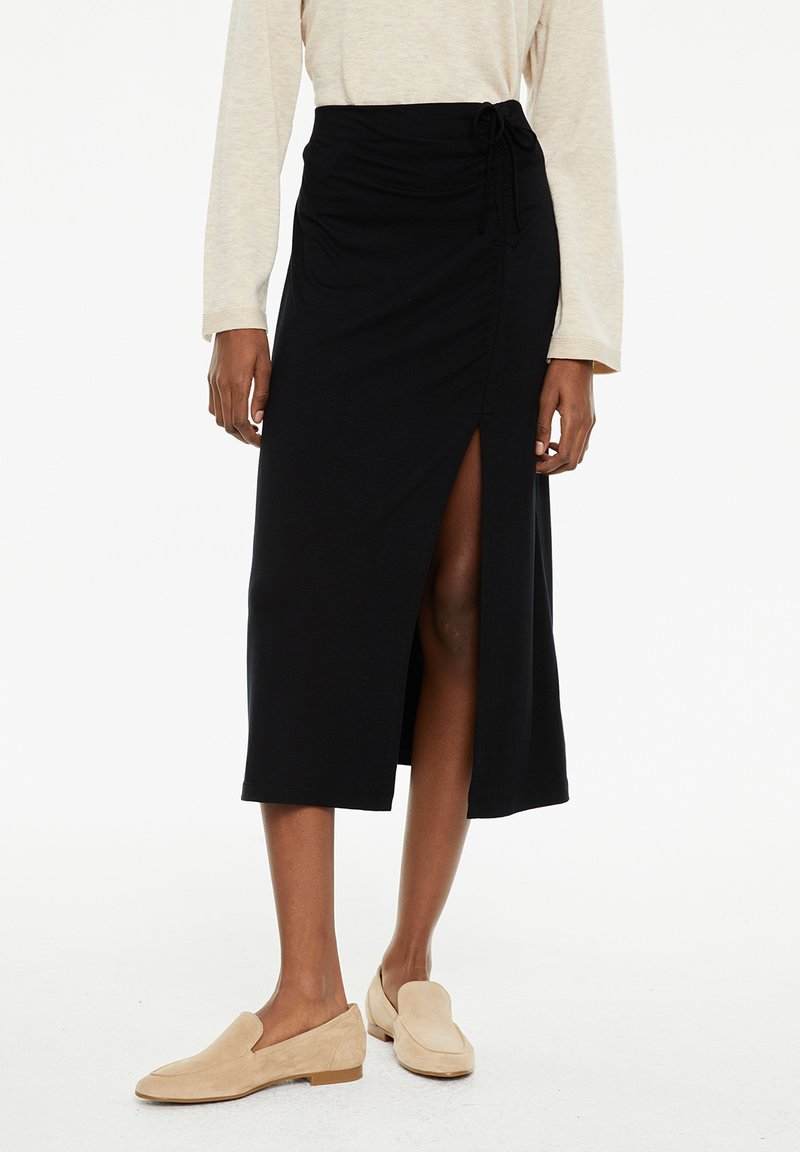 Falda midi negra de tela suave con una abertura frontal. Cuenta con una cintura con cordón y una textura suave, combinada con zapatos de lona ligeros en color beige claro.