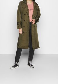Trench de color verde oliva con botones de doble botonadura, hombreras y puños anchos; llevado sobre una camisa rosa y unos vaqueros negros, combinado con zapatillas.
