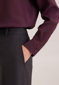 Bordeauxfarbene Bluse mit langen Ärmeln, ausgestattet mit einem Knopfverschluss am Bündchen, kombiniert mit schwarzen Hosen, die Seitentaschen und eine glatte Stofftextur aufweisen.