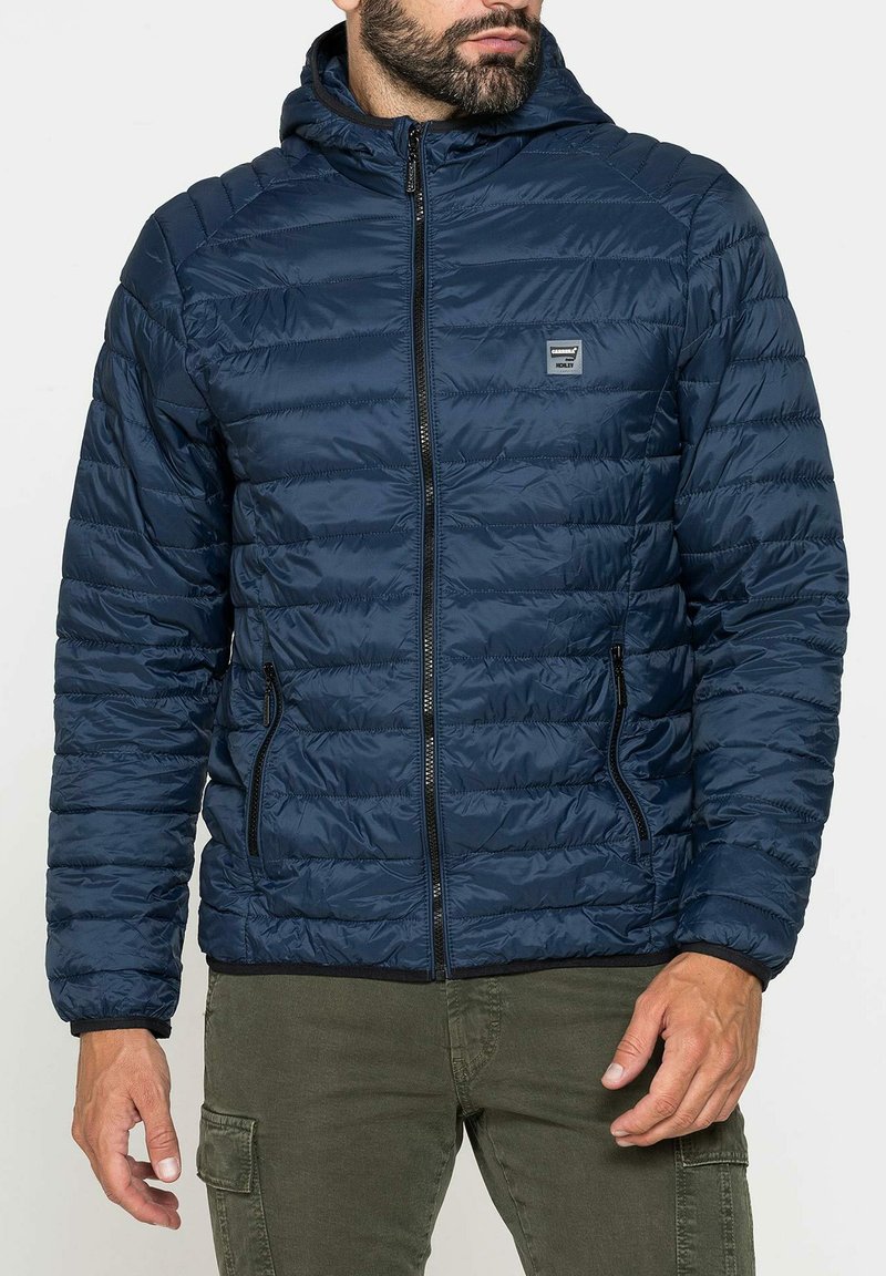Giacca imbottita blu navy con chiusura frontale a zip, cappuccio e tasche laterali. Il tessuto ha una texture trapuntata con cuciture visibili.