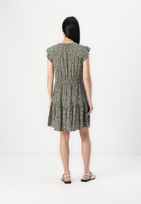 Blumenkleid in Grün mit kurzen, fließenden Ärmeln und einem gestuften Saum. Besitzt einen elastischen Bund. Getragen mit flachen Sandalen.