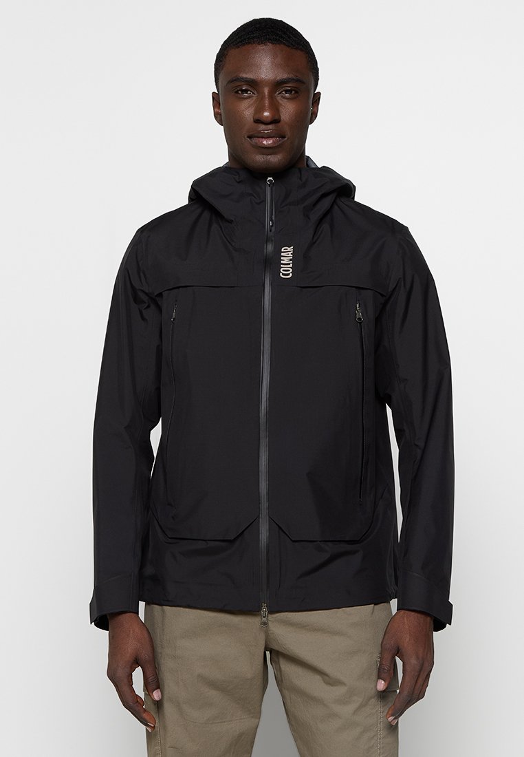 Giacca nera impermeabile con cappuccio, dotata di chiusura con zip, due tasche sul petto con zip e un logo sul lato sinistro. Texture liscia.