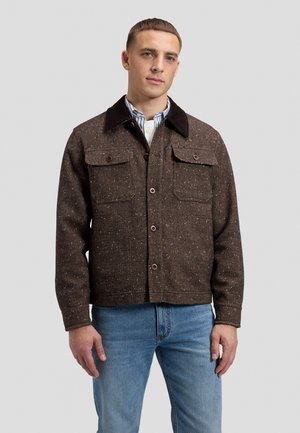 Homme portant une veste marron texturée boutonnée sur une chemise rayée et un jean bleu, debout devant un fond gris clair uni.