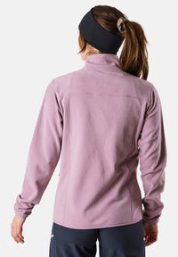 Vaaleanpunainen fleece-takki, jossa on korkea kaulus, pitkät hihat ja litteät saumat. Päällä musta hiuspanta ja hiukset poninhännällä.