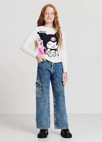 Chemise blanche à manches longues avec un graphique cartoon coloré, assortie à un pantalon cargo large en denim bleu avec poches latérales et détails tachetés.