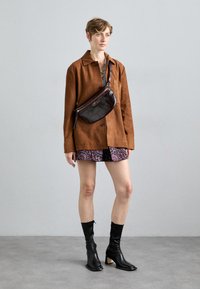 Een grote bruine suède jas, bloempatronen shorts en zwarte enkellaarsjes. Een donkere handtas wordt crossbody gedragen. Zachte texturen en een eigentijds ontwerp.