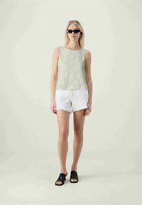 Top sem mangas de textura verde claro combinado com calções brancos de bainha desfiada e sapatos de deslizamento pretos. A modelo usa pequenos óculos de sol pretos.