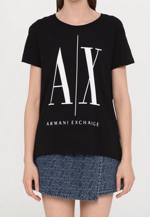 Kobieta ubrana w czarny t-shirt Armani Exchange z dużym białym logo A|X, w połączeniu z niebieską, wzorzystą dżinsową mini spódnicą.