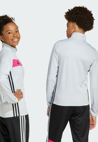 Witte sportieve jas met een roze horizontale streep en zwarte accenten. Bevat een hoge kraag en lange mouwen. Zwarte broek met drie strepen.