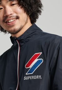 Chaqueta cortavientos azul marino con un logo bordado en rojo y azul en el pecho, cremallera blanca y diseño de cuello alto.