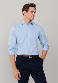 Lichtblauw overhemd met een subtiel stippenpatroon. Lange mouwen opgerold bij de manchetten. Draagt een donkere broek en een bruine riem.