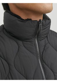 Schwarze gesteppte Jacke mit hohem Kragen, ausgestattet mit einem Reißverschluss und weicher Fleece-Fütterung am Hals. Strukturiertes Material sorgt für Tiefenwirkung.
