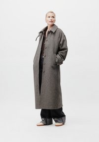 Ξανθιά γυναίκα που φοράει ένα μακρύ καρό παλτό, σκοτεινά φαρδιά τζιν με γυρισμένα μπατζάκια και λευκά αθλητικά παπούτσια, πόζαρε με τα χέρια στις τσέπες.