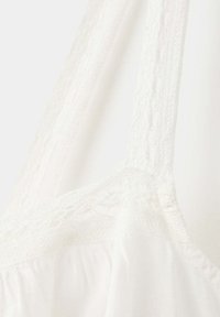 Vêtement blanc sans manches avec des bretelles en dentelle délicates et une bordure en dentelle le long de l'encolure, fabriqué dans un tissu léger.